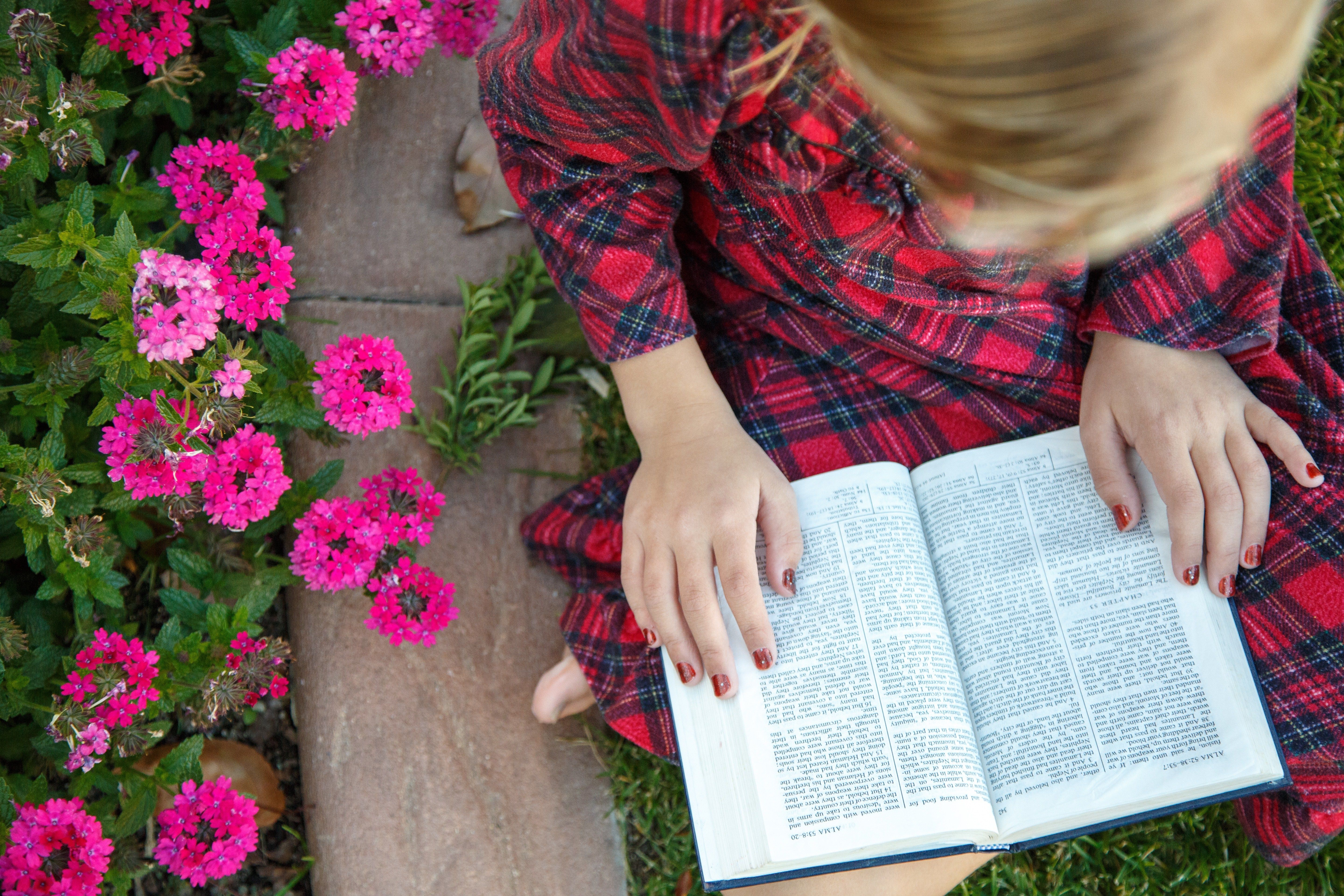 A young girl sits in a garden and reads the Book of Mormon.