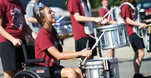 A young girl sits in a wheelchair while playing the drums. She is part of a parade