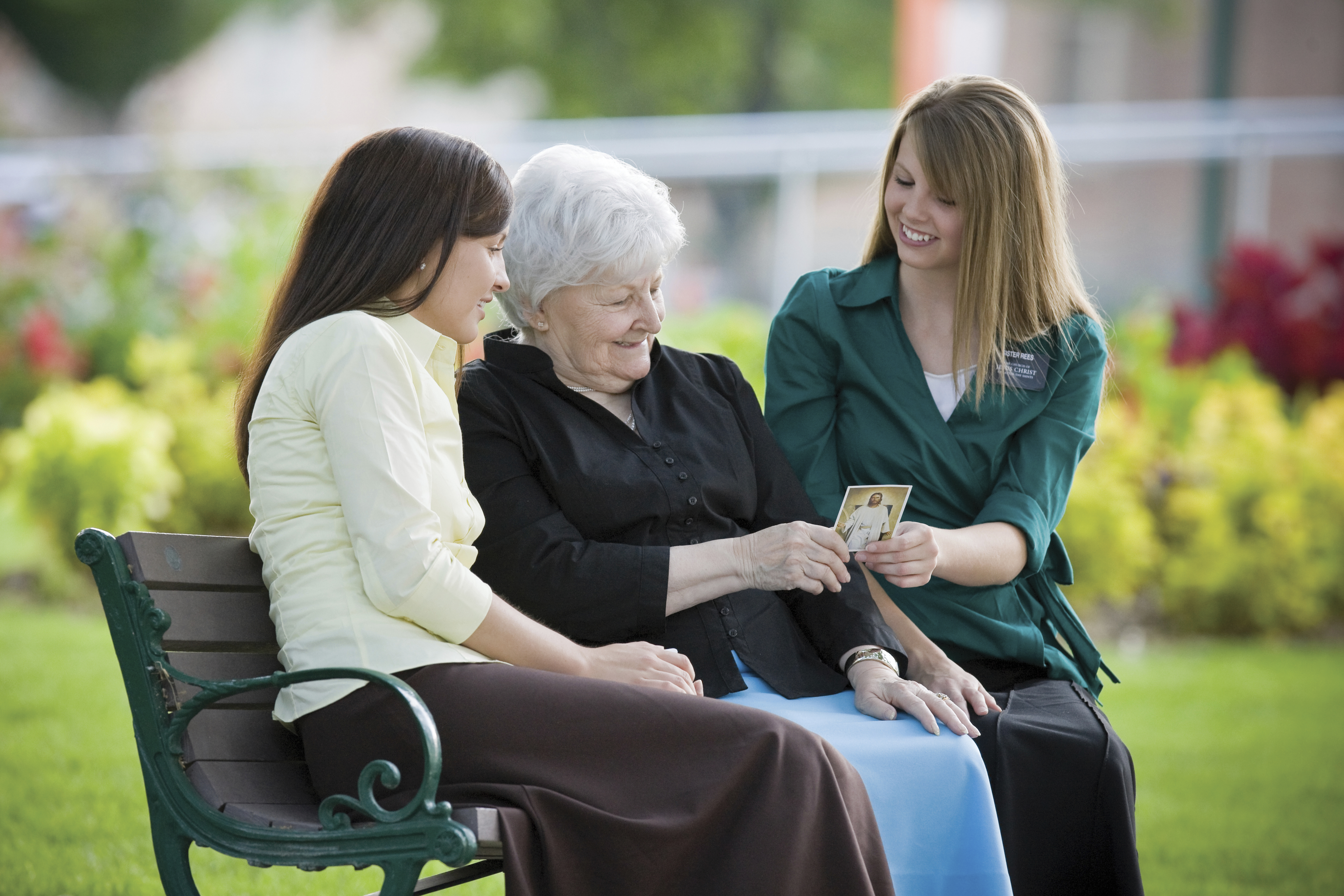 An elderly woman sitting between two sister missionaries and looking at a pass-along card.