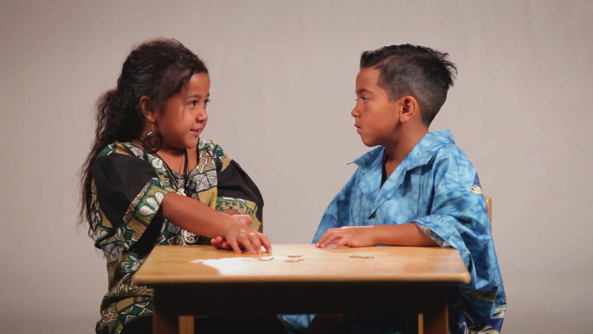 A photo of a young girl and young boy sitting at a desk, looking at each other while talking.