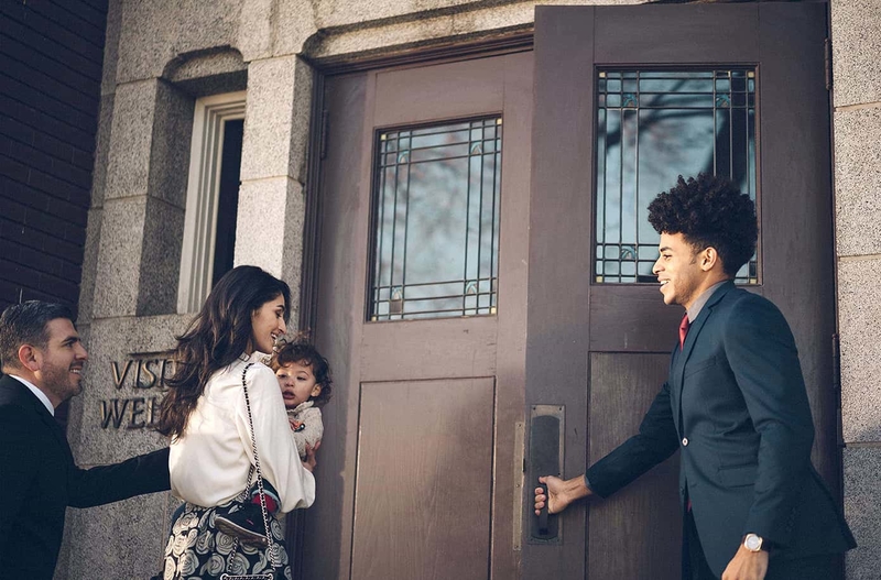 A man holds a door open for a young family into a church to learn about baptism into the church of Jesus Christ