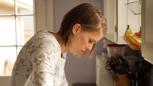 A woman is standing in her kitchen and is next to the kitchen sink. She is leaning on the sink and bowing her head in prayer.