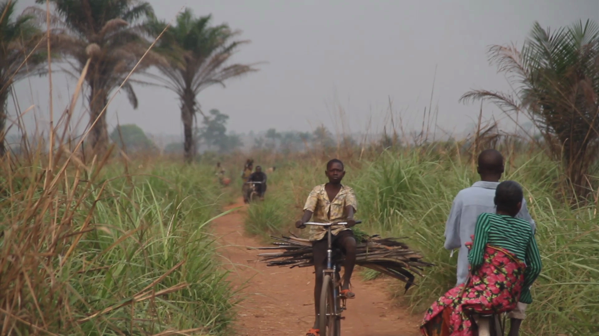 A young man riding a bike on a dirt path carrying bananas while others walk by.