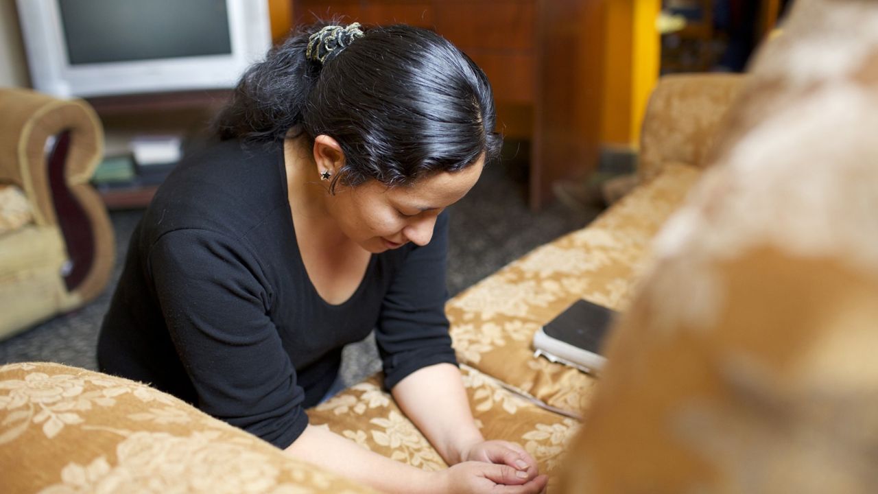 A woman kneels in prayer in her living room asking God to please help her